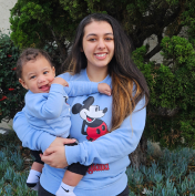 A woman holds a smiling baby outdoors. Both are wearing matching blue Mickey Mouse sweatshirts. There's greenery in the background.